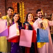 Flaunting Diwali shopping bags and greeting with smiles, Indian Asian young married couples in traditional festive attire stand at decorated home interior and show shopping bags to camera with joy
